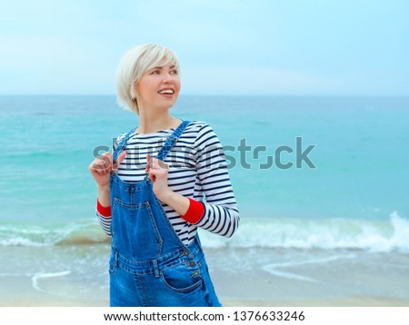 beautiful young blonde caucasian woman on vacation in striped blouse, sneakers and denim overall jumping on the beach by the amazing blue sea background