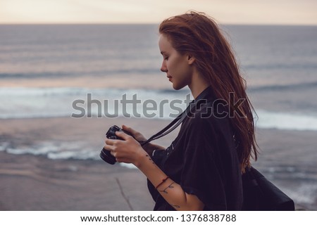 Beauty woman make photo on the ocean beach,Photography and travel. Young woman in hat holding camera sitting on wooden fishing pier with beautiful tropical sea view. Bali, vintage camera,tattoo, cute