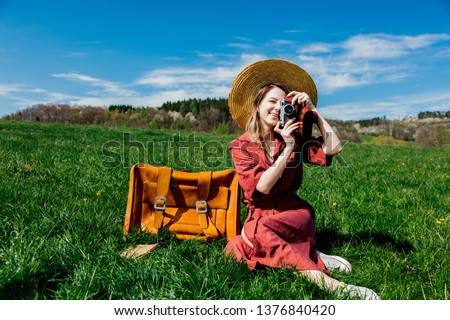 Beautiful girl in red dress and hat sitting on meadow with vintage camera and suitcase. Spring season