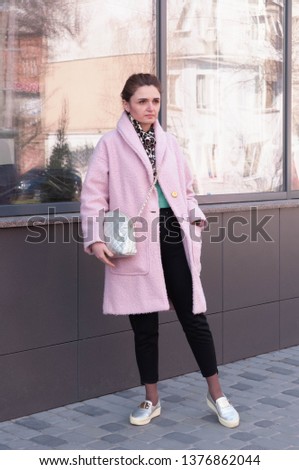 woman in pink coat and black pants standing on sidewalk, looking away near building with large windows on street