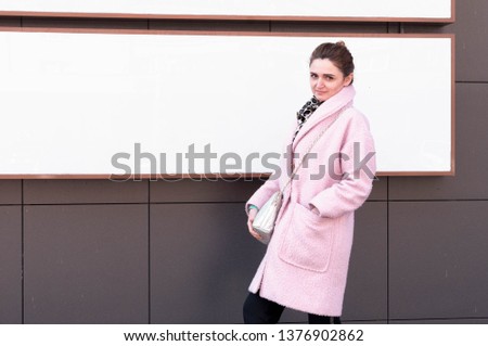 attractive woman in pink coat, black pants, bun on head standing near dark bilding with large empty board and looking at camera