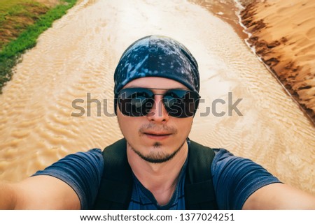 man on a hike with a backpack takes a selfie on camera face in sunglasses and a bandana while traveling on a stream background in Asia