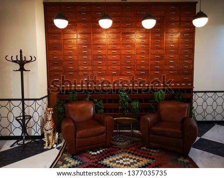 Retro home decoration consisting of 2 brown-leather armchairs, a diamond-patterned rug, a small metal table, a panther mascot, a hanger, and a large vintage wooden organizer full of tiny drawers.