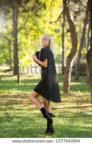 Outdoor fashion portrait of young beautiful blonde woman wearing dark dress posing in summer green park holding a hat and looking into camera smiling