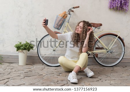 Girl sits on the ground next the bicycle and smiling during take a selfie on her smartphone - Image