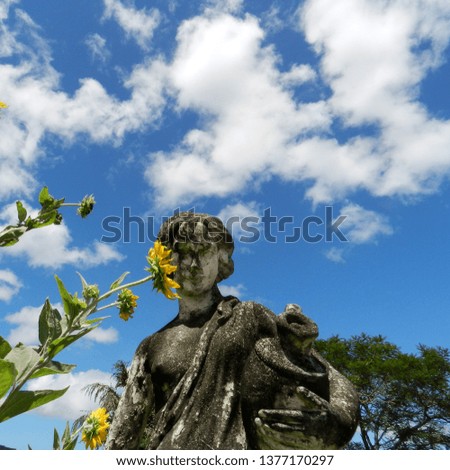 Close up a garden statue covered with mosses, yellow sunflowers. In the background the blue sky with white clouds.