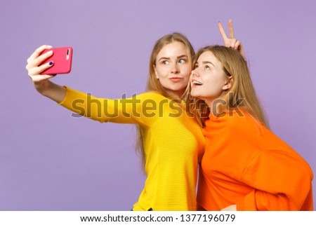 Two crazy young blonde twins sisters girls in colorful clothes doing selfie shot on mobile phone isolated on pastel violet blue wall background. People family lifestyle concept. Mock up copy space