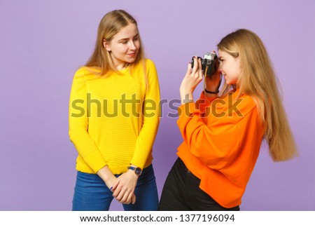 Two smiling young blonde twins sisters girls in colorful clothes taking pictures on retro vintage photo camera isolated on violet blue background. People family lifestyle concept. Mock up copy space