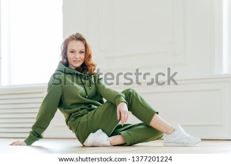 Sideways shot of satisfied redhead woman has makeup, wears tracksuit, white spotshoes, rests on floor, does stretching exercises, is fond of sport. People, flexibility and motivation concept Royalty-Free Stock Photo #1377201224