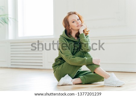 Staying in good shape. Pleasant looking red haired female gymnast with wavy hair, sits on floor in fitness hall, has flexible body, rests after doing yoga exercises, looks confidently at camera Royalty-Free Stock Photo #1377201239