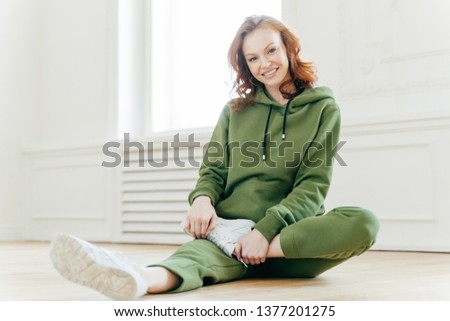 Photo of happy red haired woman in sweatsuit, white sneakers, takes rest after workout, sits on floor, stretches legs, has active healthy lifestyle. Smiling fitness girl does exercising. Flexibility