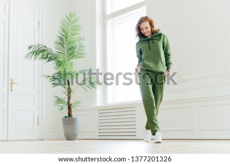Full length shot of active pleased young woman with slim body, keeps hands in pockets of tracksuit, focused down, ready for doing cardio exercises, has determined look, pilates exercising indoor Royalty-Free Stock Photo #1377201326