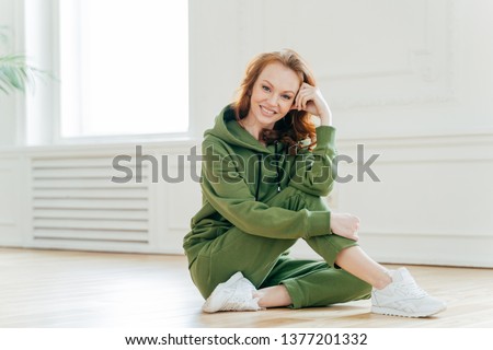 Positive redhead fitness girl practices healthy lifestyle, has rest after indoor workout, feels excited after active exercises, wears comfortable green sweatsuit, poses on floor. Physical activity Royalty-Free Stock Photo #1377201332