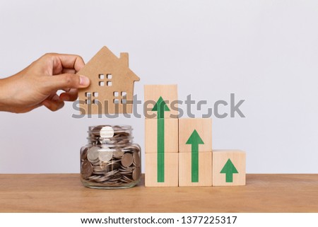 Hand putting house model on money coins in glass jar withWooden cube stack a staircase and arrows pointing up. Real estate investment concept