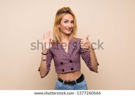 Young blonde woman with pink jacket over isolated wall showing ok sign and thumb up gesture