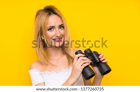 Young blonde woman over isolated yellow wall with black binoculars