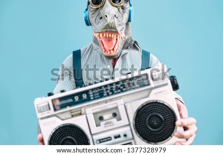 Crazy senior man wearing t-rex mask and listening to music holding vintage boombox stereo outdoor - Fashion masquerade male having fun dancing and celebrating outside - Absurd and funny people concept