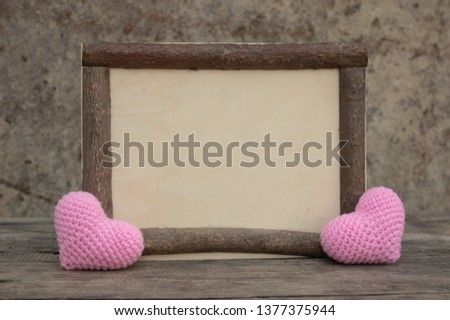 Brown wooden frame on the rough wooden table with pink knitting hearts. Background of the rock wall. Sun light shines on the frame. Copy space for edting and text.