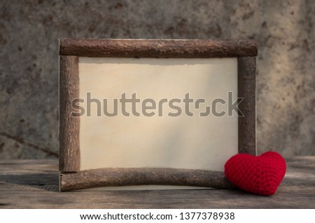 Brown wooden frame on the rough wooden table with the red knitting heart on the corner. Background of the rock wall. Sun light shines on the frame. Copy space for edting and text.