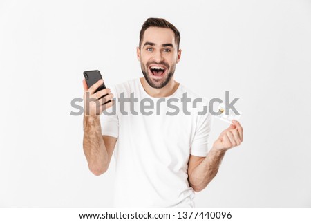 Handsome cheerful man wearing blank t-shirt standing isolated over white background, using mobile phone, showing plastic credit card