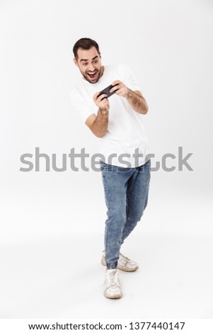 Full length of a handsome cheerful man wearing blank t-shirt standing isolated over white background, playing games on mobile phone