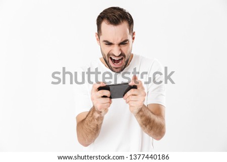 Handsome angry man wearing blank t-shirt standing isolated over white background, playing games on mobile phone