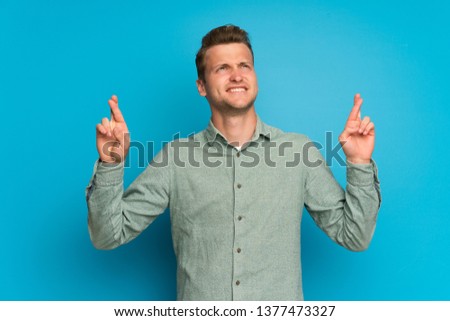 Blonde man over isolated blue wall with fingers crossing and wishing the best