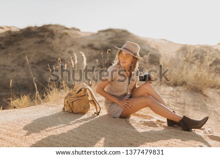 beautiful stylish young woman in khaki dress in desert traveling in Africa on safari wearing hat and backpack taking photo on vintage camera, exploring nature, hot summer, traveler on vacation