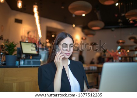 Confident female executive director reading summary on laptop computer and phoning to new specialist while sitting in restaurant during work break. Woman talking via mobile phone during distance job. 