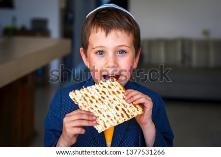 Cute Caucasian child in a yarmulke taking a bite from a traditional Jewish matzo unleavened bread in a room. Royalty-Free Stock Photo #1377531266