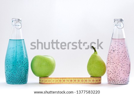 A centimeter, an apple, a pear and glass bottles with pink and blue basil seed stand on a white background.