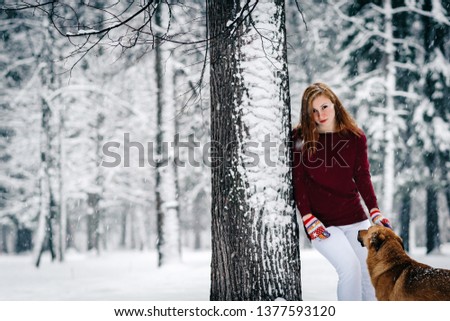 a beautiful girl in a Burgundy sweater and white pants stands leaning against a tree near the Red Dog amid the snowcovered forest