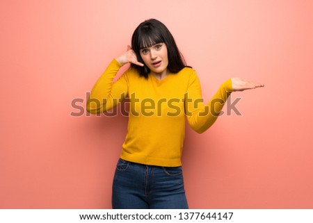 Woman with yellow sweater over pink wall making phone gesture and doubting