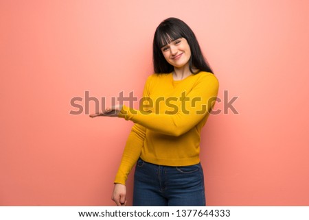 Woman with yellow sweater over pink wall presenting an idea while looking smiling towards