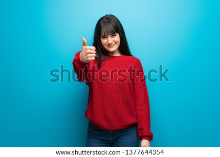 Woman with red sweater over blue wall with thumbs up because something good has happened