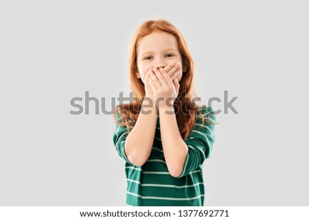 childhood, embarrassment and emotions concept - confused red haired girl in green striped shirt covering her mouth by hands over grey background