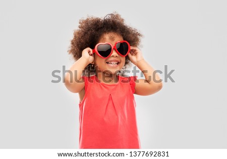 childhood, valentine's day and summer concept - happy little african american girl in heart shaped sunglasses over grey background Royalty-Free Stock Photo #1377692831