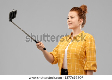 technology and people concept - smiling red haired teenage girl in checkered shirt taking picture by smartphone on selfie stick over grey background