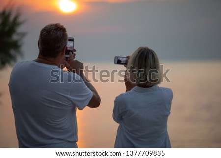 A man and a woman taking pictures of the sea sunset on their phones, we see them from the back