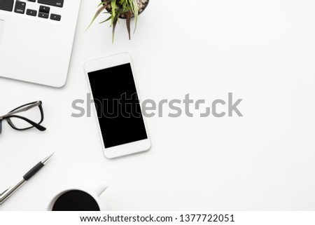White smartphone with black blank mock up screen is on top of white office desk table with supplies. Top view with copy space, flat lay.