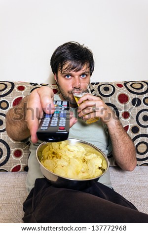 Young man watching television, eating potato chips and drinking beer indoor