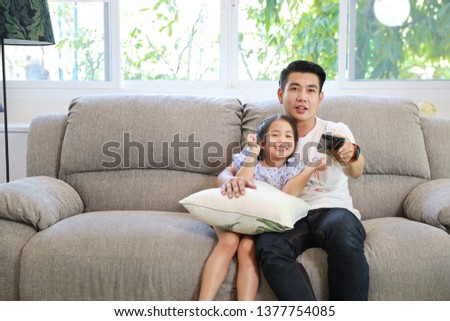 Happy Asian family, father and daughter are watching TV on the sofa in the living room and he is pressing the remote with a smiling face (relaxation and technology concept)