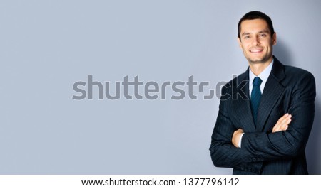 Portrait picture of confident businessman in black suit and blue tie, with crossed arms pose, empty copy space place for some text, advertising or slogan, over grey background. Business concept photo.