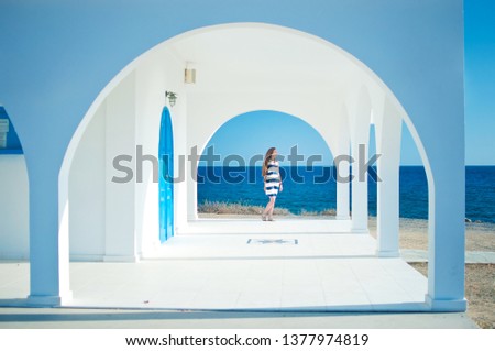 Image of caucassian girl in striped dress with long brown hair looking in the distance, against blue sea. Warm summer day. White arch and columns of Ayia Thekla Chapel