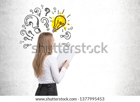 Portrait of young blonde businesswoman with copybook looking at concrete wall with question marks and lightbulb drawn on it. Concept of idea. Mock up