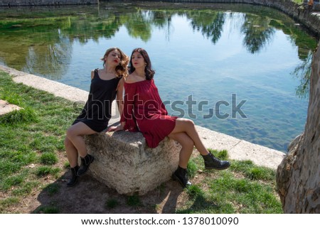 Romantic picture of two female friends along the shores of a lake. Sitting on a stone bench, a blonde in a black dress, the other brunette in a red dress. Concept of relaxing outdoors.