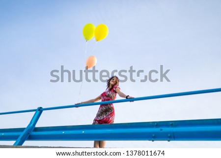 Young beautiful girl with red dress, boots and helium balloons is happy on a jetty by the ocean.