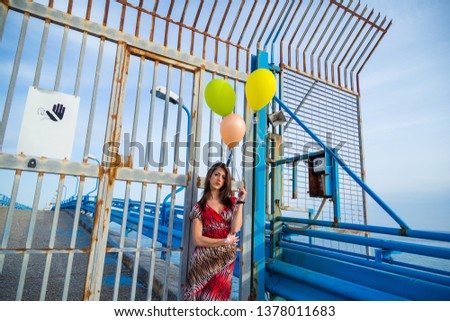 Young beautiful girl with red dress, boots and helium balloons is happy on a jetty by the ocean.