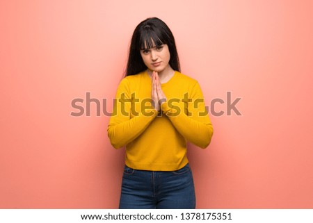 Woman with yellow sweater over pink wall keeps palm together. Person asks for something
