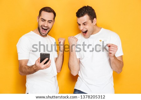 Two cheerful excited men friends wearing blank t-shirts standing isolated over yellow background, looking at mobile phone, celebrating success